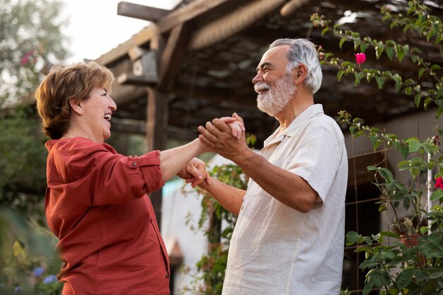 Smiling senior couple dancing together in a garden, holding hands and enjoying time outdoors near flowering plants.