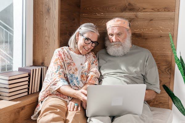 A senior couple sitting together indoors using a laptop, smiling while browsing online in a cozy wooden room.