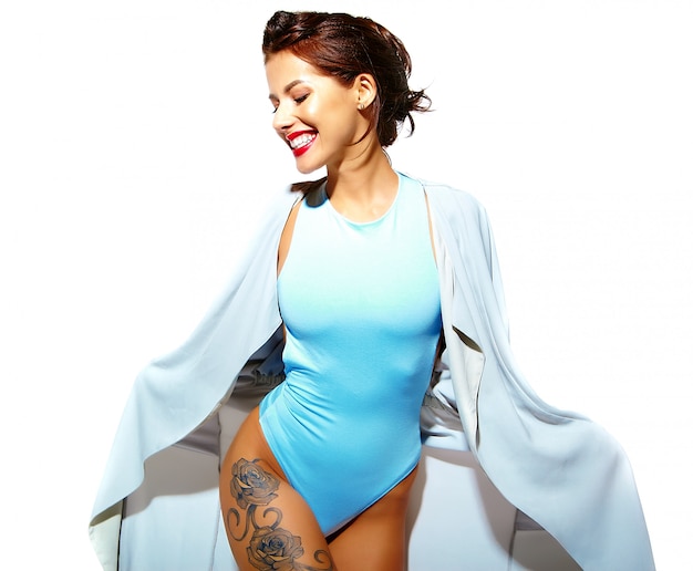 Smiling woman in blue swimsuit and light robe showing leg tattoos on white background.
