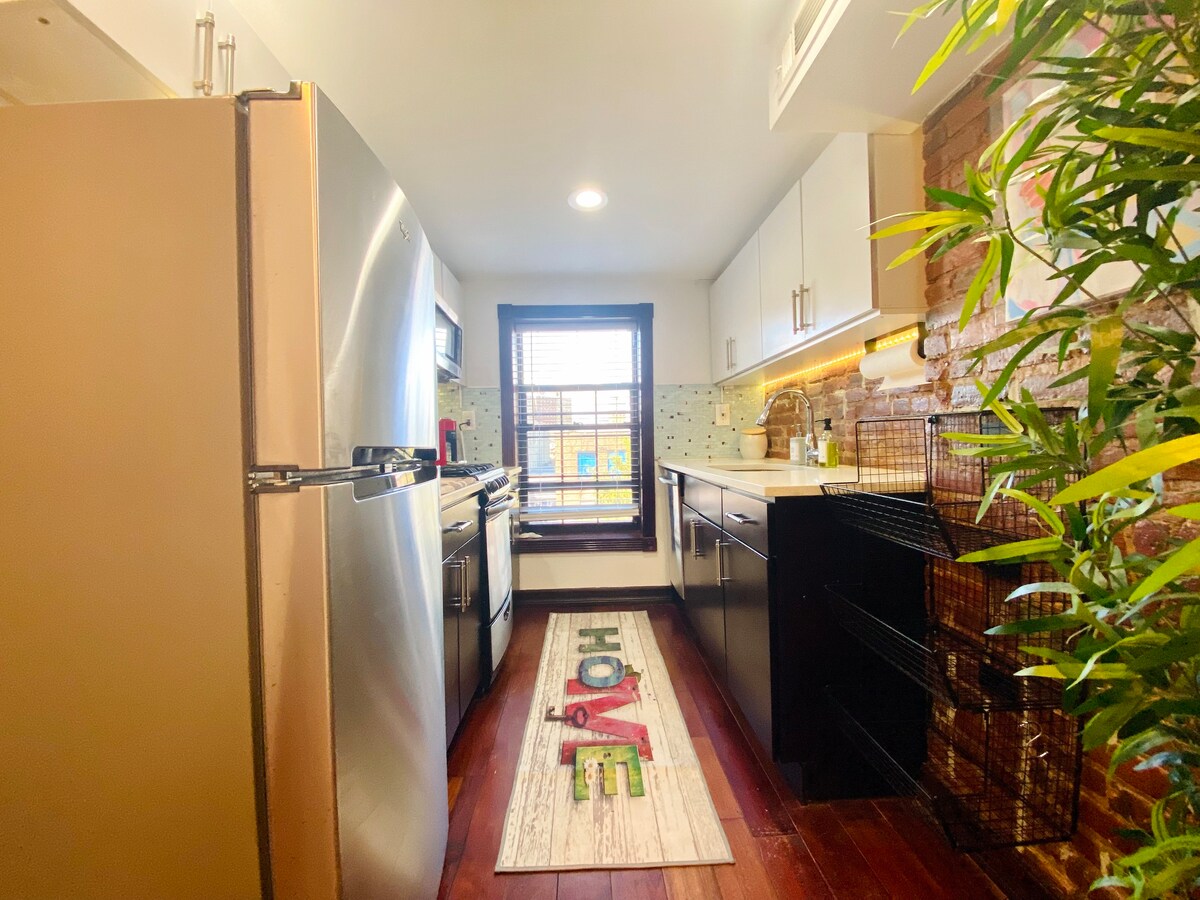A cozy kitchen with wooden floors, a stainless steel refrigerator, dark cabinets, and a window above the sink. A colorful "HOME" rug lies on the floor, and a potted plant adds greenery to the space.