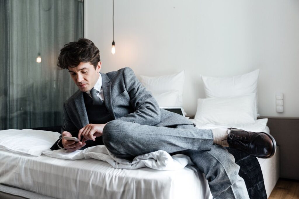 A man in a gray suit sits casually on a bed, holding a smartphone. He is wearing black shoes and socks, with one leg crossed over the other. The room has a modern, minimalist design with soft lighting and neutral tones.