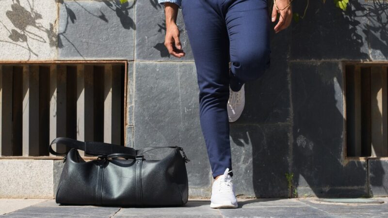 A person in a stylish outfit—navy blue pants, white sneakers, and a striped shirt—is walking outdoors. They carry a sleek black duffel bag on their shoulder, with one leg raised mid-step against a textured urban backdrop.