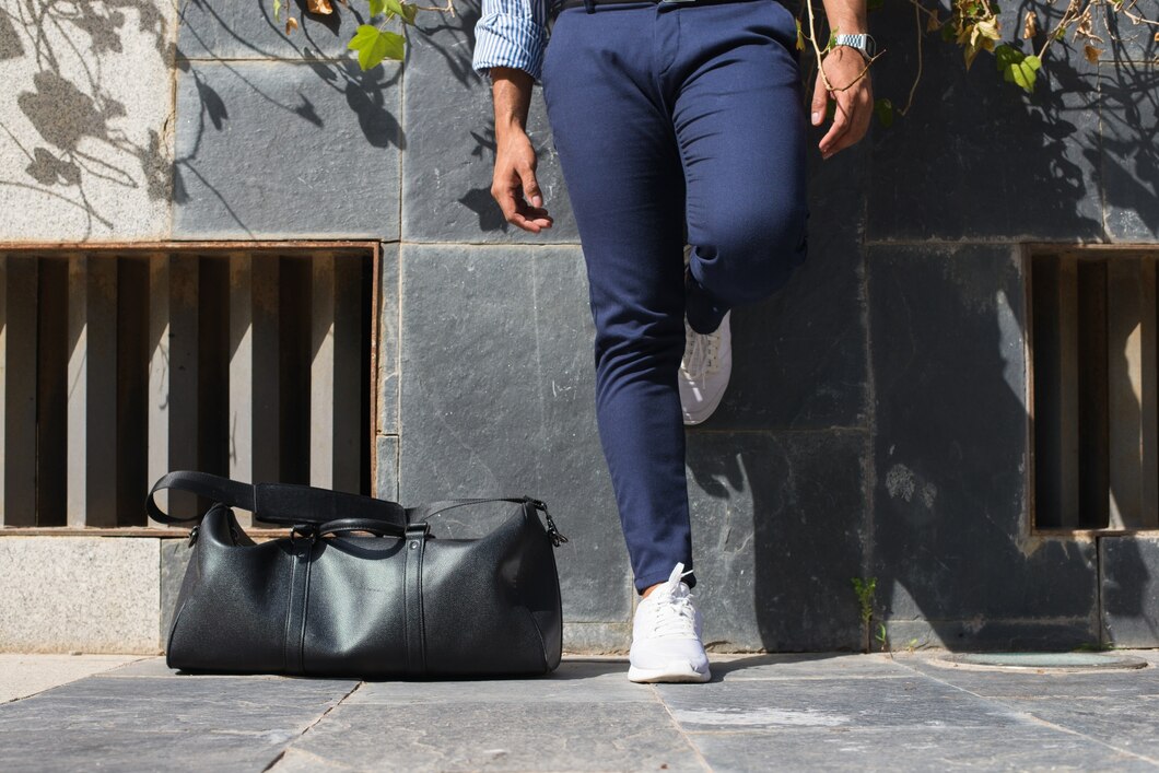 A person in a stylish outfit—navy blue pants, white sneakers, and a striped shirt—is walking outdoors. They carry a sleek black duffel bag on their shoulder, with one leg raised mid-step against a textured urban backdrop.