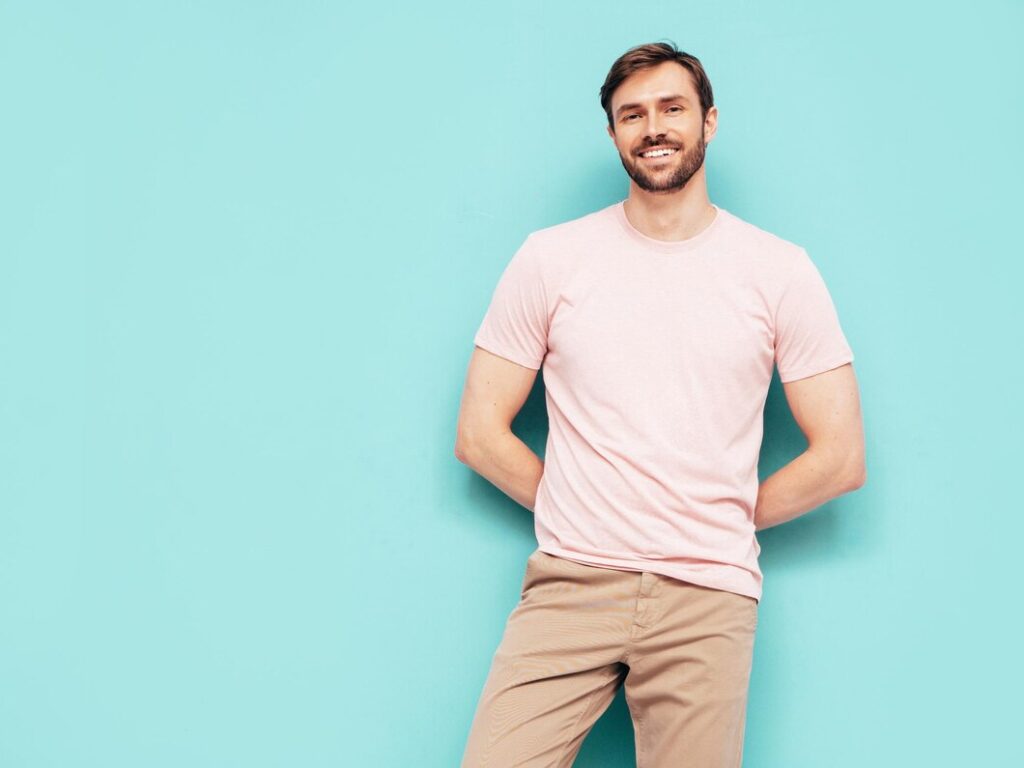 A man stands confidently against a light blue background, wearing a pink t-shirt and beige pants. He has a warm smile and is looking directly at the camera.