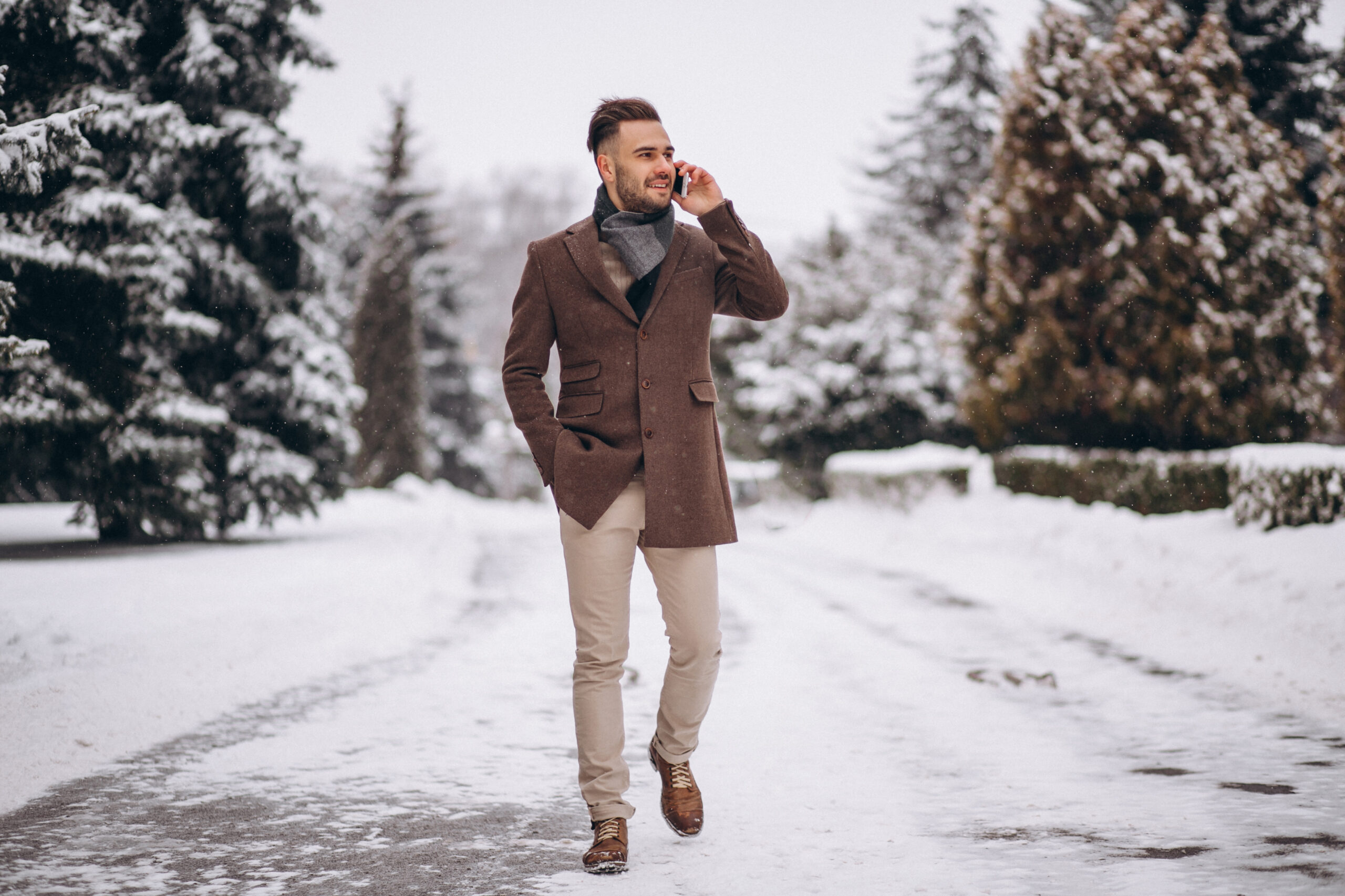 A man walks on a snowy path while talking on his phone, dressed in a brown coat, scarf, beige pants, and boots amid winter trees.