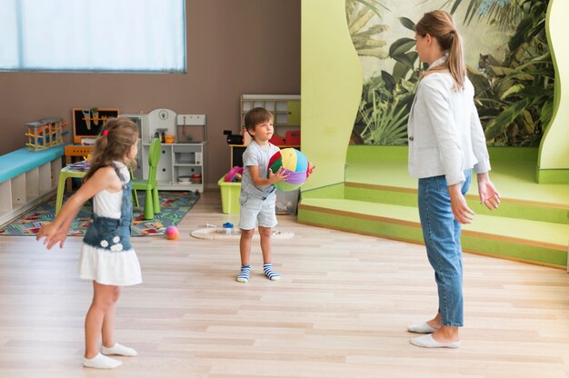 A teacher leading two young children playing with a ball in a bright preschool classroom.