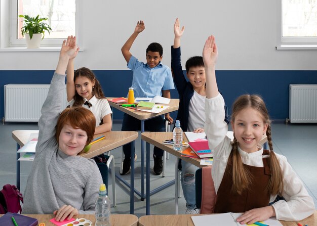 Diverse group of elementary school students sitting at desks, smiling and raising their hands to answer a question.